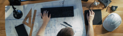 View of a man at his desk. On his desk are two computer monitors and an architect's blueprint.
