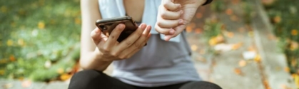 Showing an example of healthcare consumerism, a woman is sting in athletic clothes. She is holding her cellphone in one hand and checking her fitness tracker in the other.