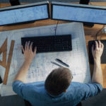 View of a man at his desk. On his desk are two computer monitors and an architect's blueprint.
