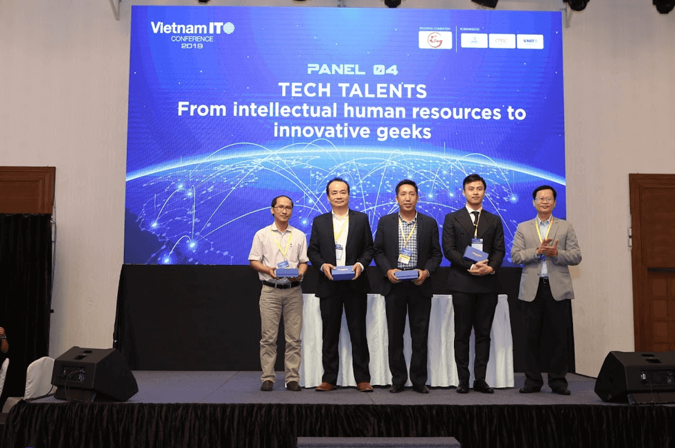 5 men stand on stage in suits. Behind them is a table with a white tablecloth and a projector screen that says "Tech Talents. From intellectual human resources to innovative geeks."
