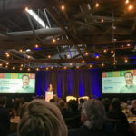This image shows the award ceremony for the Leaders in Corporate Citizenship Award. A man stands on stage behind a podium addressing the crowd. To his left and right are two screens with Josh's photo and the KMS logo.