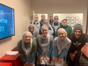 Three women stand smiling, wearing white plastic aprons, blue plastic arm covers, and hairnets. The two on the right are giving a thumbs up.