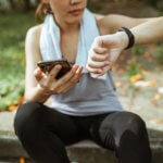 Showing an example of healthcare consumerism, a woman is sting in athletic clothes. She is holding her cellphone in one hand and checking her fitness tracker in the other.